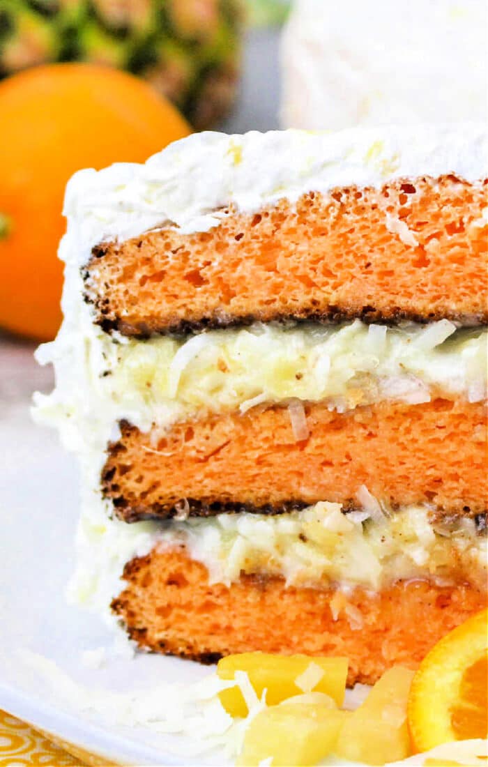 A close-up of a slice of layered orange and white Hawaiian cake with frosting, featuring visible layers of orange cake, white frosting, and shredded coconut. There is an orange and some pineapple pieces in the background.