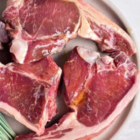 Three raw lamb chops elegantly arranged on a round beige plate, inviting you to ponder how long to cook lamb chops for the perfect tenderness.