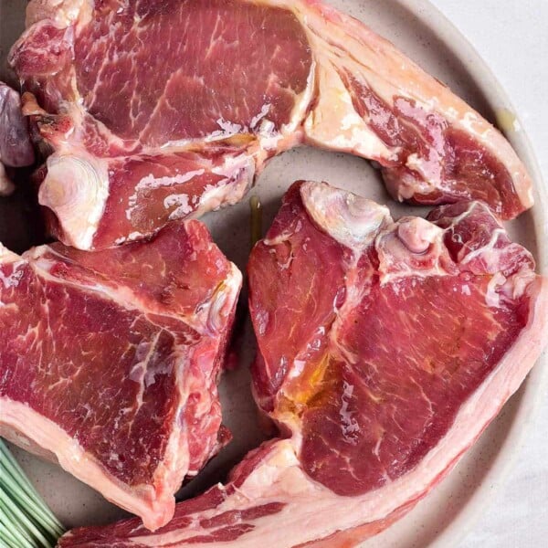 Three raw lamb chops elegantly arranged on a round beige plate, inviting you to ponder how long to cook lamb chops for the perfect tenderness.