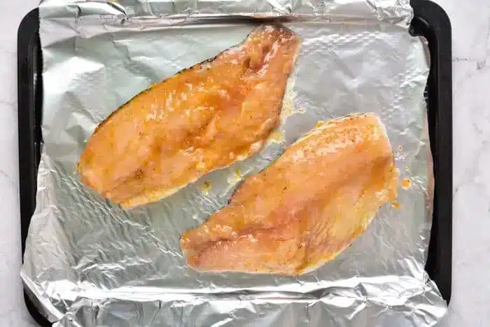 Two raw fish fillets, perfectly sauced, are placed on a foil-lined baking tray, ready to transform into a delightful meal as they bake from frozen.