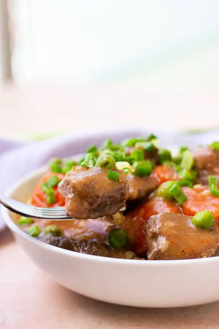 A close-up of a bowl of savory stew with chunky pieces of bear meat, carrots, and green peas. A fork holds a tender piece above the bowl, while the dish is beautifully garnished with chopped green onions.