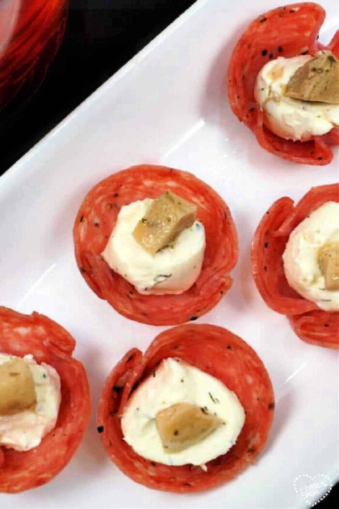 A close-up of a white rectangular plate showcasing an appetizer. The salami cups feature a creamy cheese mixture filling, topped with a small piece of artichoke. Five salami cups are arranged delicately on the plate.