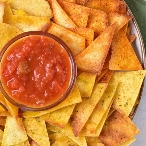 A plate of yellow and orange tortilla chips, made using an air fryer for a crispy texture, surrounds a bowl of red salsa. The plate rests on a surface where a green cloth peeks out from underneath.