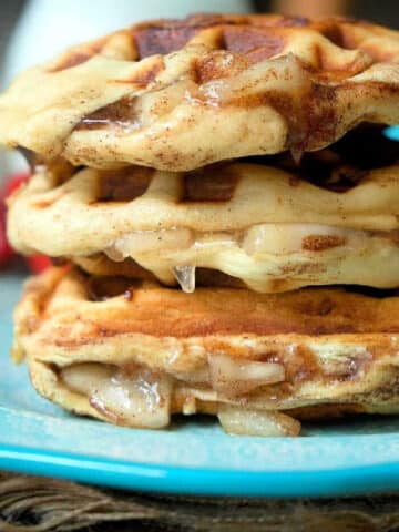 Close-up of a stack of three glazed, apple pie stuffed waffles on a blue plate. The waffles show a golden-brown, slightly crispy texture with a dripping glaze. The background is blurred, putting focus on the cinnamon roll waffles. Text 