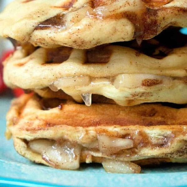 Close-up of a stack of three glazed, apple pie stuffed waffles on a blue plate. The waffles show a golden-brown, slightly crispy texture with a dripping glaze. The background is blurred, putting focus on the cinnamon roll waffles. Text "The Typical Mom" is faintly visible at the bottom right.