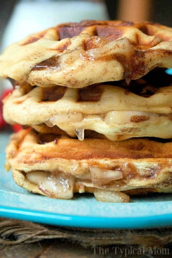 Close-up of a stack of three glazed, apple pie stuffed waffles on a blue plate. The waffles show a golden-brown, slightly crispy texture with a dripping glaze. The background is blurred, putting focus on the cinnamon roll waffles. Text "The Typical Mom" is faintly visible at the bottom right.
