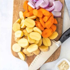 Chopped potatoes, carrots, and red onions on a wooden cutting board await a knife's touch; spices in small bowls nearby enhance the flavor of steaming vegetables in a microwave.