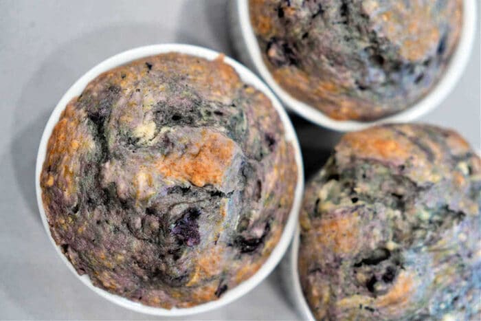 A close-up image of three Cake Mix Blueberry Muffins. The muffins have a golden-brown crust with visible blue and purple specks from the blueberries. They are in white paper muffin cups and are placed on a light-colored surface.