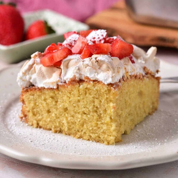 A slice of sponge cake topped with meringue, diced strawberries, and powdered sugar sits on a white plate