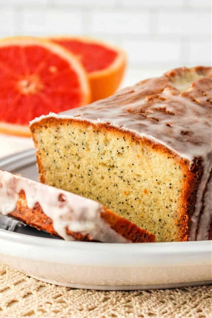 A loaf of poppy seed bread with a glaze sits on a white plate. A slice has been cut from the loaf and is placed next to it, resembling a grapefruit cake. In the background, there are two halves of a grapefruit. The scene is set on a textured beige surface.