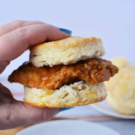 A hand grasps a delicious Whataburger Honey Butter Chicken Biscuit, with its golden layers tempting the eye, while a plate and another biscuit wait invitingly in the background.