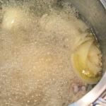 A pot of boiling water with pieces of meat and sliced onions sits on a stovetop. The water is actively bubbling, showing it's at a rolling boil. At the top, the text reads, "How to Boil Meat.