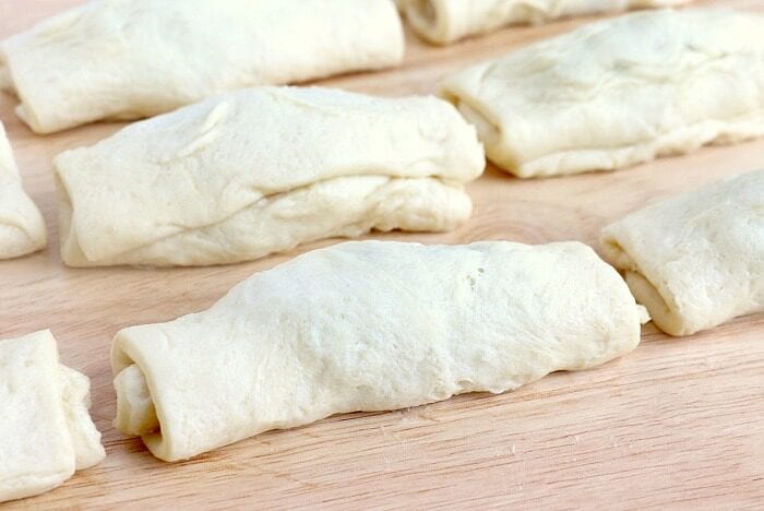 Several unbaked pastry rolls are placed on a wooden surface. The dough is light in color with a slightly textured appearance. The rolls, reminiscent of peanut butter and jelly donuts, are arranged in an uneven pattern, ready for baking.