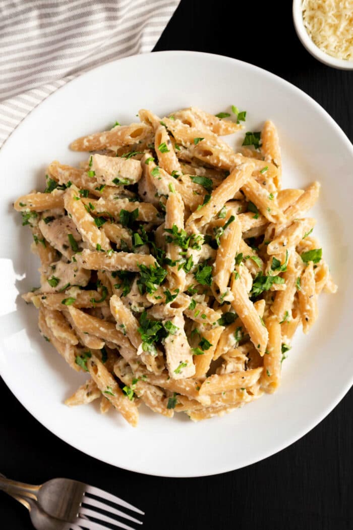 A white plate filled with creamy penne pasta garnished with chopped fresh herbs. The pasta is mixed with a rich, cheesy sauce reminiscent of slow cooker chicken alfredo. In the background, there is a small bowl of grated cheese, part of a cloth, and a fork on a dark surface.