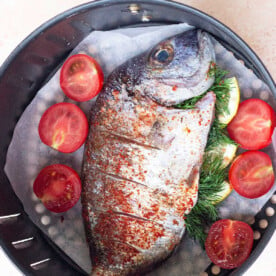 Air fryer whole fish seasoned with herbs and layered with sliced tomatoes rests in the basket, ready for cooking perfection.