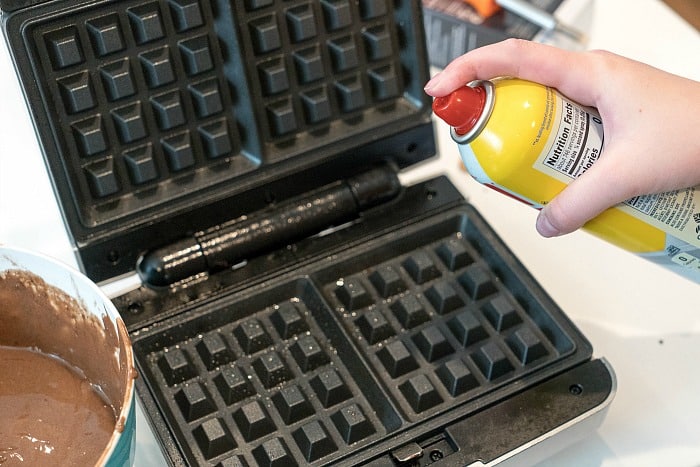 A person sprays cooking oil on a waffle iron next to a bowl of batter, getting ready to master the art of making waffles with cinnamon rolls.