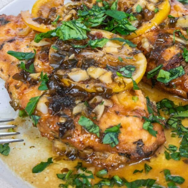 Grilled chicken topped with lemon slices and chopped herbs rests elegantly on a white plate, embodying the zest of Crockpot Chicken enhanced with Italian Dressing.