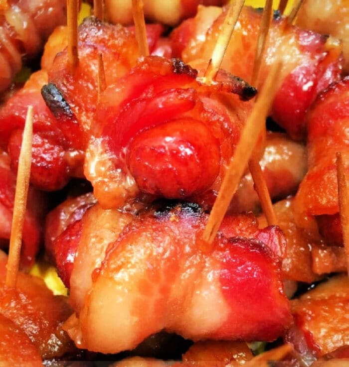 Close-up image of Bacon Wrapped Smokies secured with toothpicks. The bacon is cooked to a crispy, golden-brown finish in the Ninja Foodi. The appetizers are closely packed, showcasing the texture and color variation from the caramelized and well-cooked bacon.