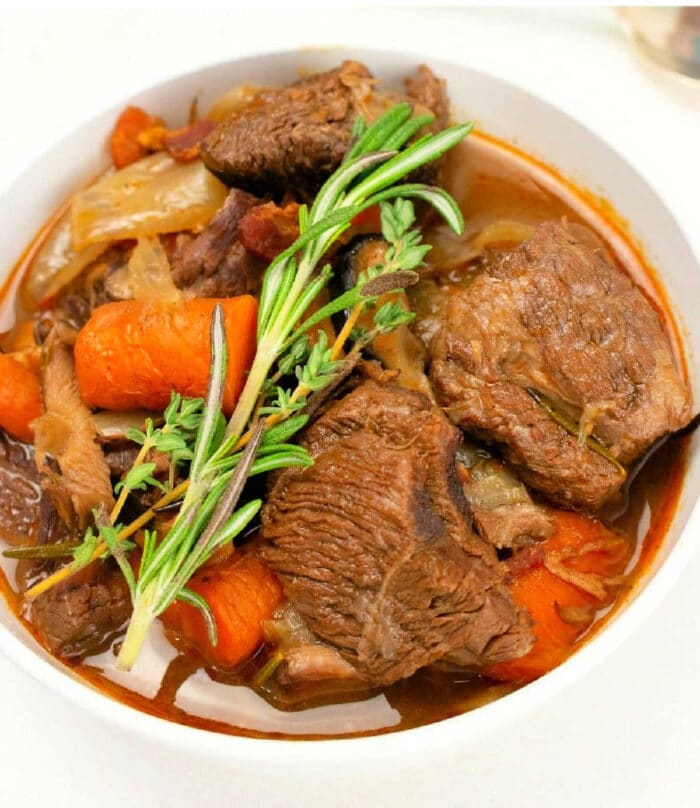 A bowl of Instant Pot Beef Bourguignon containing large chunks of beef, carrots, celery, and onions, garnished with sprigs of fresh rosemary and thyme in a rich broth. The dish is served in a white bowl on a light surface.