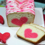 A surprise cake loaf with pink heart shapes inside and pink and white icing on top, served on a white board.
