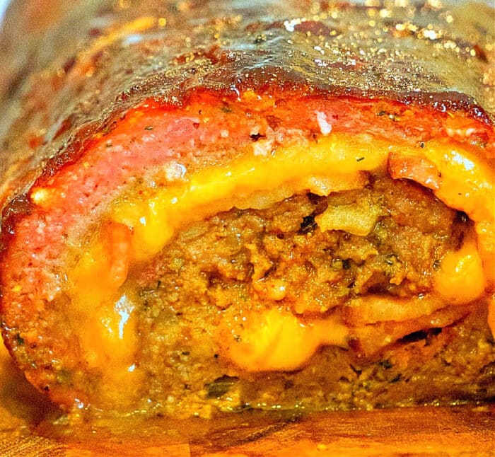 Close-up of a cross-section of a smoked meatloaf roll. This tempting smoked meatloaf recipe is filled with melted cheese and ground meat, all wrapped in a layer of bacon. The outer layer is glazed and browned, giving it a delightful caramelized appearance.