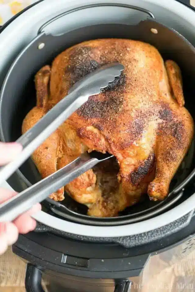 A seasoned cooked whole chicken is being held by metal tongs inside a pressure cooker. The chicken, reminiscent of one prepared as an air fryer whole chicken, appears to be browned and crispy on the outside.