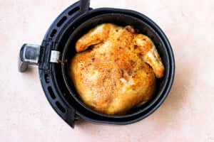 In an air fryer basket, a whole chicken with browned, crispy skin rests on a light pink textured surface.