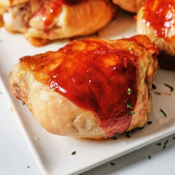 BBQ chicken thighs with a glossy glaze, garnished with herbs, served on a white square plate. Oven-baked to perfection.