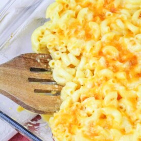 A clear glass baking dish filled with copycat Chick Fil A mac and cheese, topped with melted and lightly browned cheese. A wooden slotted spatula rests in the dish, partially lifting a portion of the macaroni. A red and white striped cloth is underneath the dish.