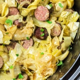 Sliced sausages with cabbage and noodles, reminiscent of a classic Haluski recipe, garnished with chopped parsley in a crockpot.