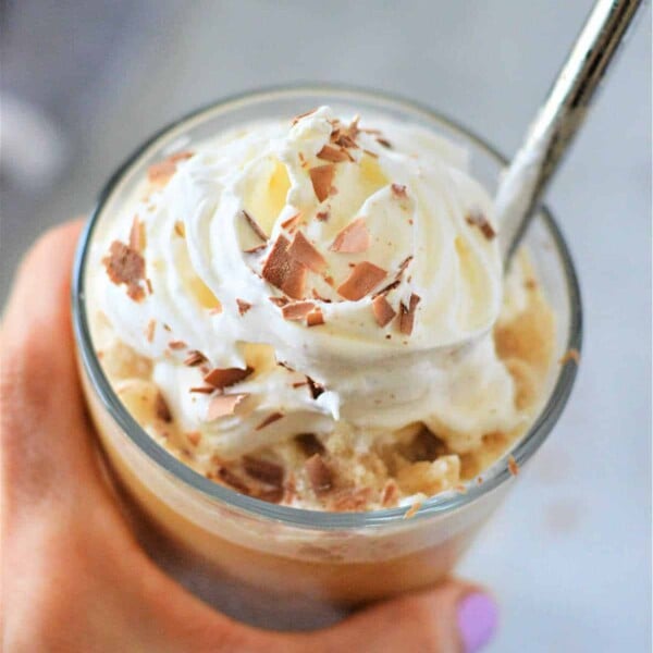 Close-up of a hand holding a frozen iced coffee topped with whipped cream and chocolate shavings, with a spoon.