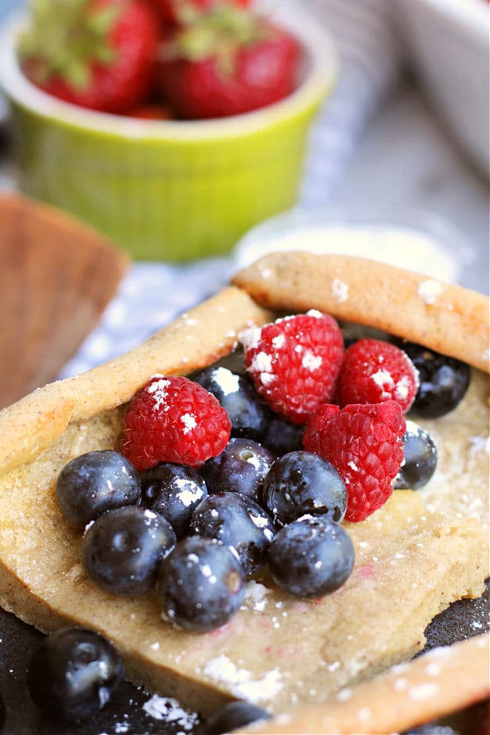 A close-up of a piece of pastry topped with powdered sugar, blueberries, and raspberries. In the background, there is a green bowl filled with fresh strawberries. The scene resembles a delightful setting where pancakes in the oven could be baking to perfection for a delicious dessert or snack.