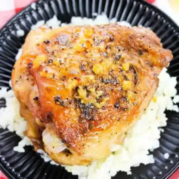 A piece of cooked chicken thigh seasoned with herbs and garlic sits on a bed of white rice on a black plate, raising the question of how long to bake bone-in chicken thighs at 350 degrees for perfect tenderness. The plate rests on a red and white checkered cloth.