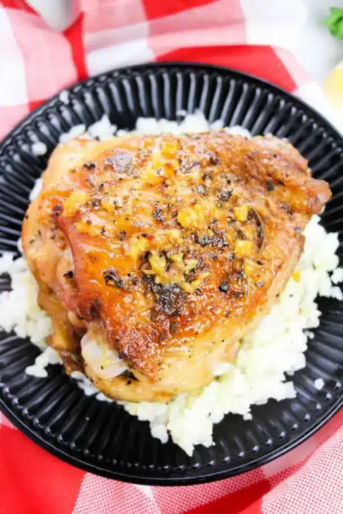 A piece of cooked chicken thigh seasoned with herbs and garlic sits on a bed of white rice on a black plate, raising the question of how long to bake bone-in chicken thighs at 350 degrees for perfect tenderness. The plate rests on a red and white checkered cloth.