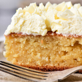 Close-up of a slice of moist lemon curd cake topped with whipped cream, placed on a plate with a fork nearby.