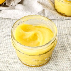 A small glass jar filled with smooth yellow lemon curd rests invitingly on a textured beige surface, enticing thoughts of delectable lemon curd desserts.