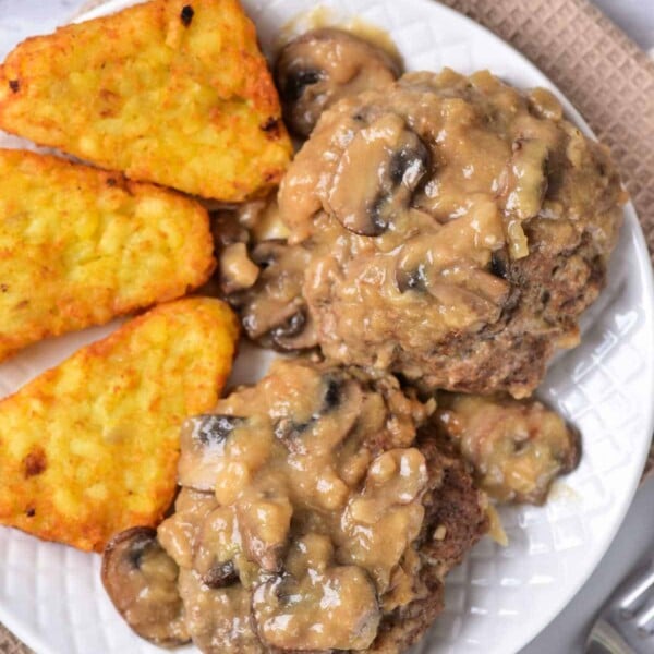 A plate showcasing an old-fashioned Salisbury steak recipe, served with rich mushroom gravy and crispy hash browns, rests elegantly on a beige placemat.