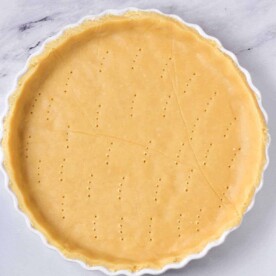 Unbaked pie crust in a white tart pan, pierced with fork holes, rests on a marble surface, ready for the next step in learning how to blind bake a pie crust.