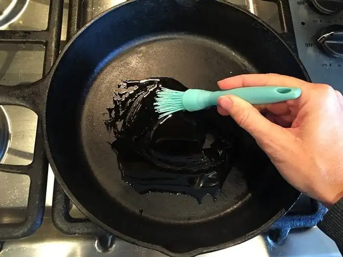 A hand holding a blue silicone brush is spreading oil on a black cast-iron skillet, preparing it for cooking. The skillet is placed on a stove with metal burners and control knobs visible on the side. Meanwhile, wondering how long to bake bone-in chicken thighs at 350 can ensure perfectly cooked results.