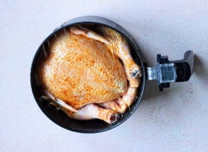 A whole chicken seasoned with spices is placed inside an air fryer basket, showcasing the simplicity and flavor of cooking an air fryer whole chicken on a light-colored countertop.