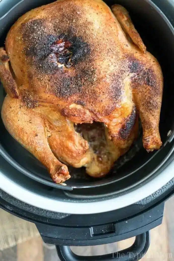 A cooked air fryer whole chicken with a seasoned, browned exterior sits in an air fryer basket. The black air fryer is placed on a wooden surface. The chicken appears to be well-roasted with crispy skin.