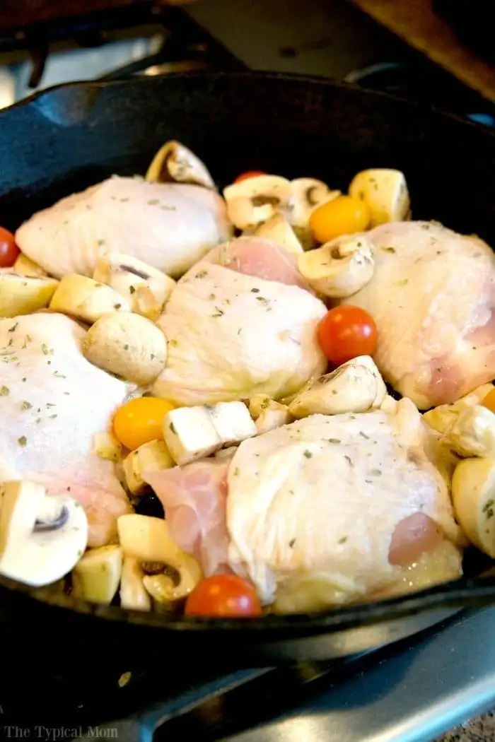 A cast iron skillet on a stove contains raw chicken thighs, quartered mushrooms, and cherry tomatoes, ready for cooking. While you prep your ingredients, you might wonder, *How long to bake bone-in chicken thighs at 350*? Seasoned with herbs, this dish promises a flavorful experience.