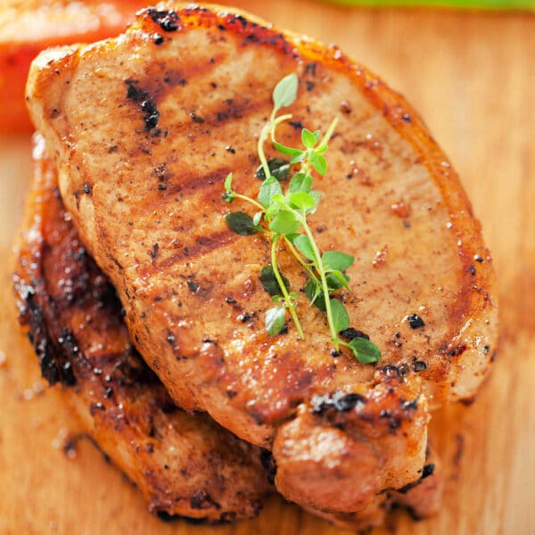 Two grilled pork chops, prepared from frozen pork chops in the air fryer, garnished with fresh herbs on a wooden board, with peppers in the background.