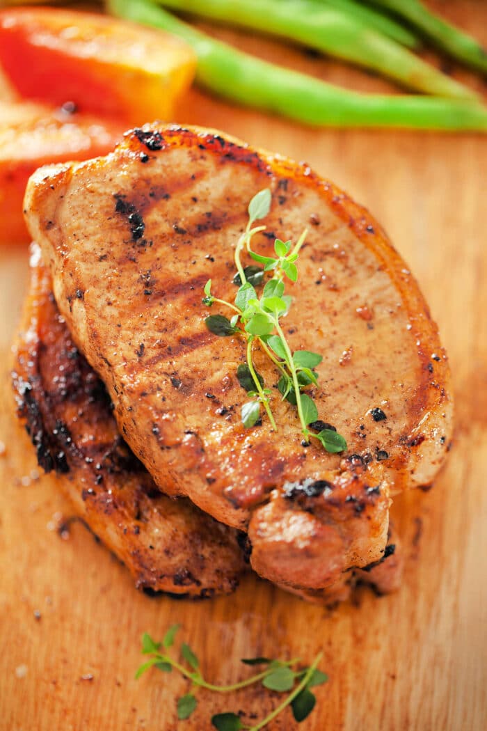 Two grilled pork chops, prepared from frozen pork chops in the air fryer, garnished with fresh herbs on a wooden board, with peppers in the background.
