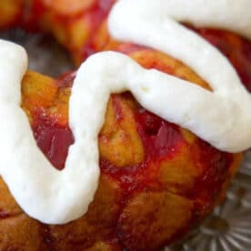 A cherry-filled bundt cake topped with a thick swirl of white icing sits on a glass plate, reminiscent of the gooey sweetness found in cinnamon roll monkey bread.