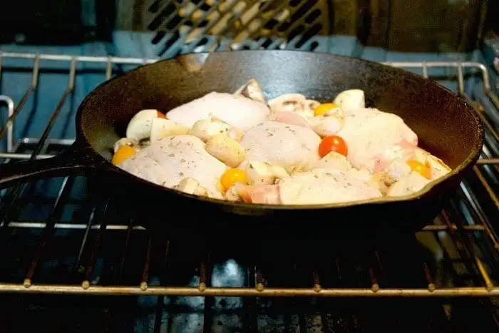 A cast iron skillet containing raw chicken pieces, cherry tomatoes, and mushrooms is in an oven with the door closed. The oven's heating elements are visible, and the food appears to be seasoned with herbs. For those wondering how long to bake bone-in chicken thighs at 350, this setup offers a savory answer.