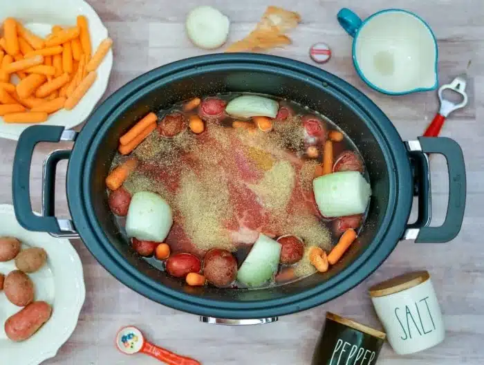 A slow cooker filled with meat, carrots, potatoes, onions, and broth sits on a table with veggies and seasonings&mdash;perfect for making a roast with onion soup mix in crockpot for a flavorful meal.