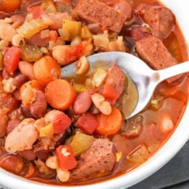 A bowl of instant pot 15 bean soup with sausage, carrots, and tomatoes, with a spoon scooping up a hearty portion.