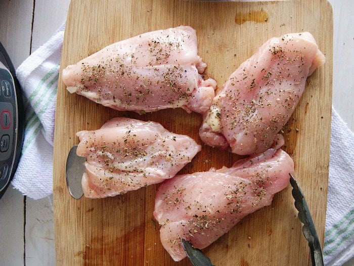 Four seasoned raw chicken thighs, perfect for a crockpot lemon pepper chicken recipe, rest on a wooden cutting board alongside a trusty pair of tongs.