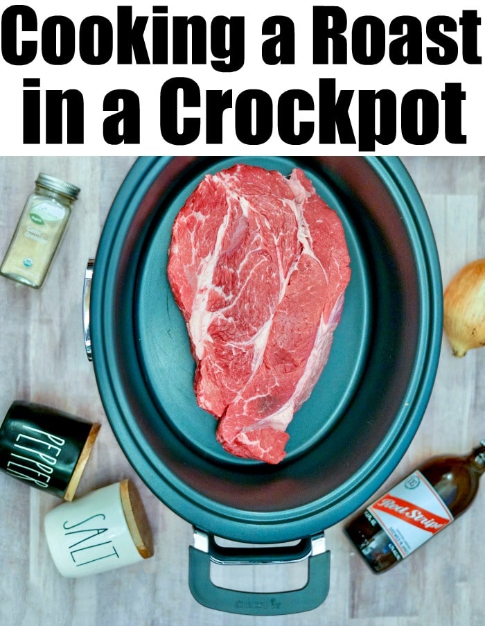 Raw beef roast in a crockpot surrounded by seasonings, an onion, a bottle of beer, and a packet of onion soup mix on a wooden surface. Perfect for an easy roast with onion soup mix in crockpot recipe.